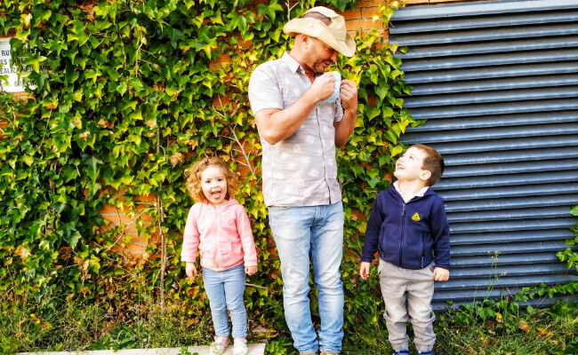 A man in jeans and a shirt wearing a white hat laughing and smiling with 2 young children